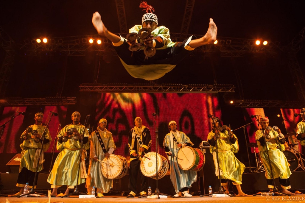 A Gnawa musician mid-jump during a high-energy performance on stage